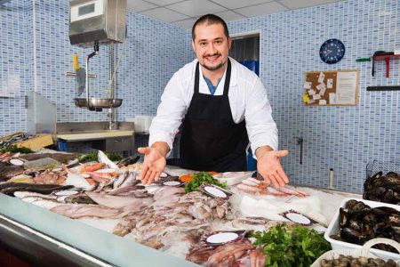Glad cheerful seller in black apron showing fish on his counter in supermarket